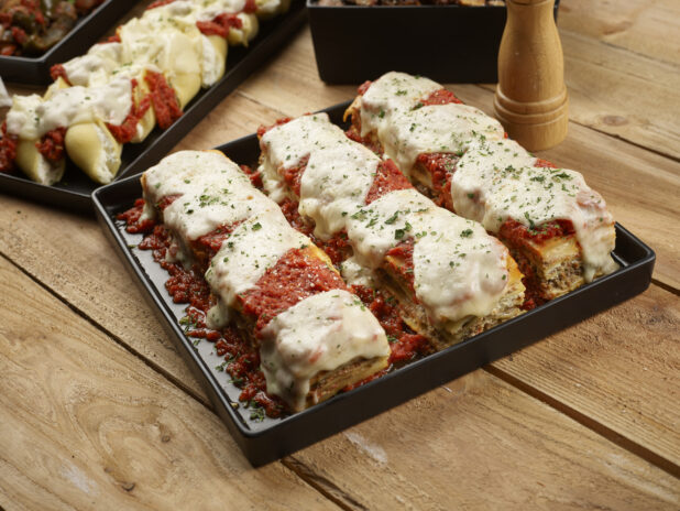 Lasagna on a black square platter with stuffed pasta shells on a rectangular black platter in the background on a wood tabletop