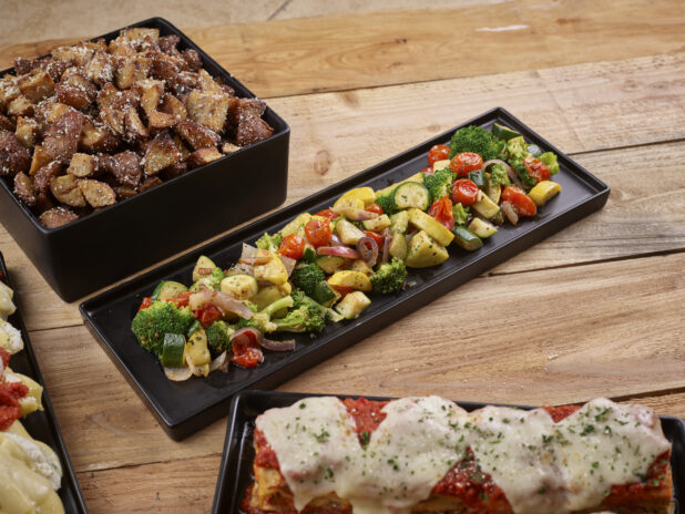 Lasagna, steamed vegetables and parmesan roasted potatoes all on rectangular and square black platters on a wooden background