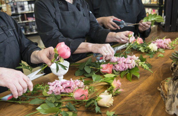 Three people trimming flowers for multiple bouquets in a line on a wooden table