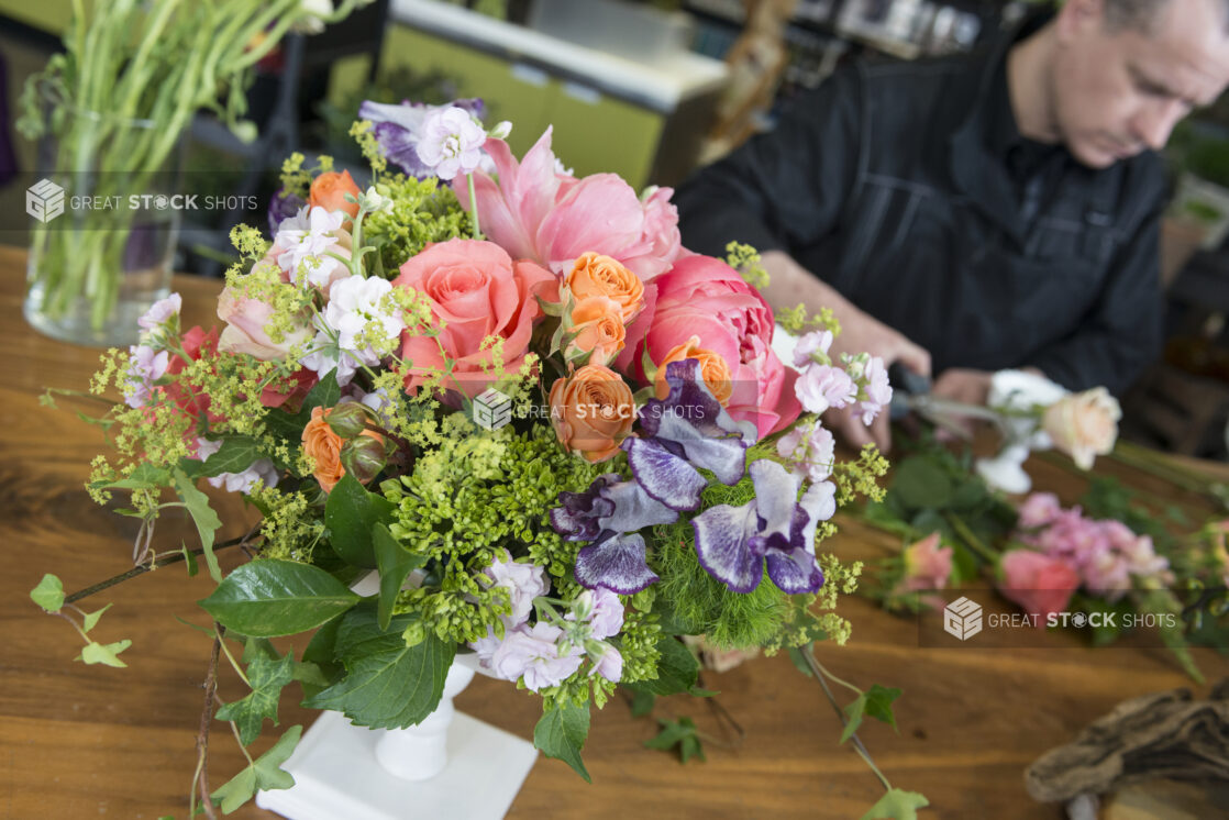 Arrangement of various flowers in a standing vase with a man trimming flowers in the background