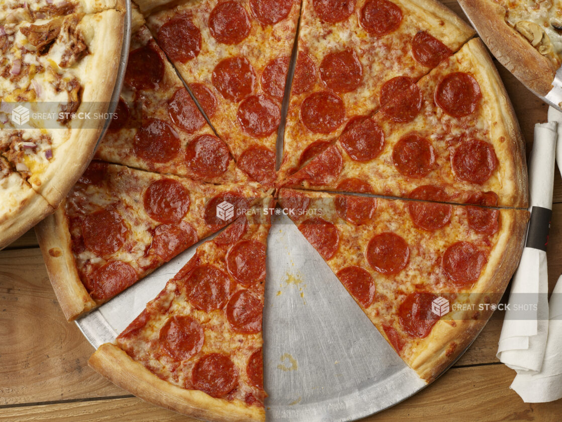 Overhead view of a sliced pepperoni pizza with rolled cutlery and pizzas on either side on a wooden background