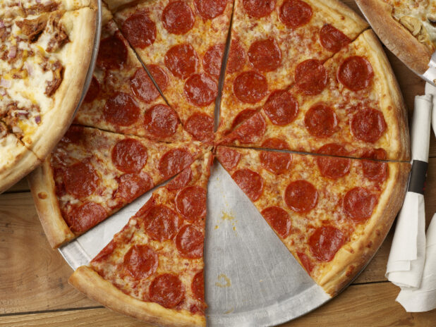 Overhead view of a sliced pepperoni pizza with rolled cutlery and pizzas on either side on a wooden background