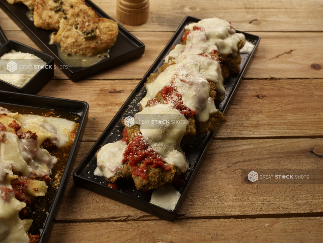 Chicken parmesan on a black rectangular platter with other Italian dishes and grated parmesan cheese surrounding it on a wooden background