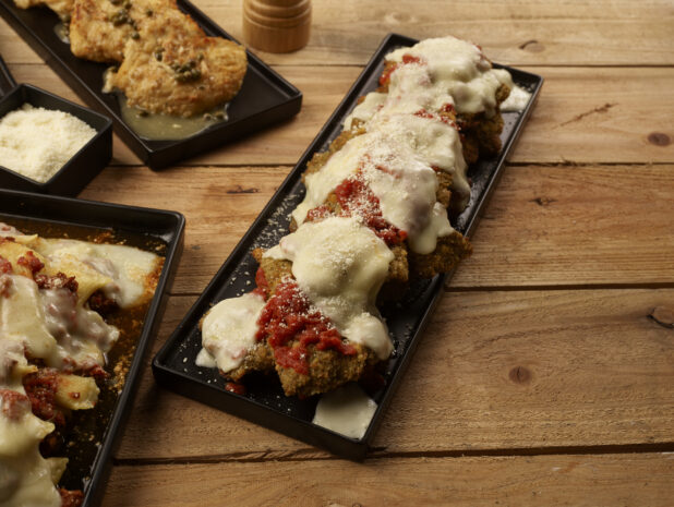 Chicken parmesan on a black rectangular platter with other Italian dishes and grated parmesan cheese surrounding it on a wooden background