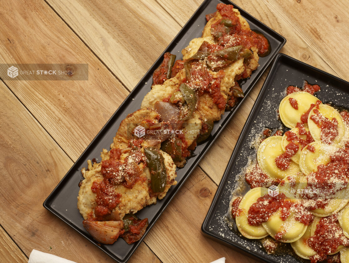 Chicken scarpariello on a rectangular black platter and mezzalune pasta (ravioli) on a square black platter on a wooden background