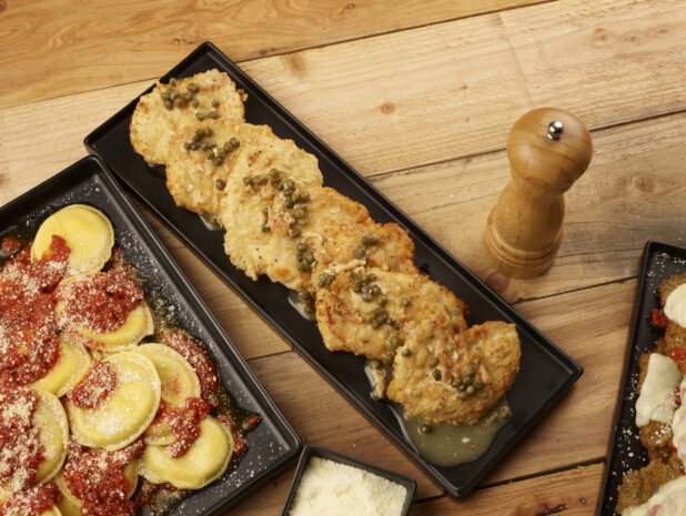 Chicken piccata and mezzalune ravioli, on rectangular and square black platters on a wooden background with a black ramekin of grated parmesan cheese