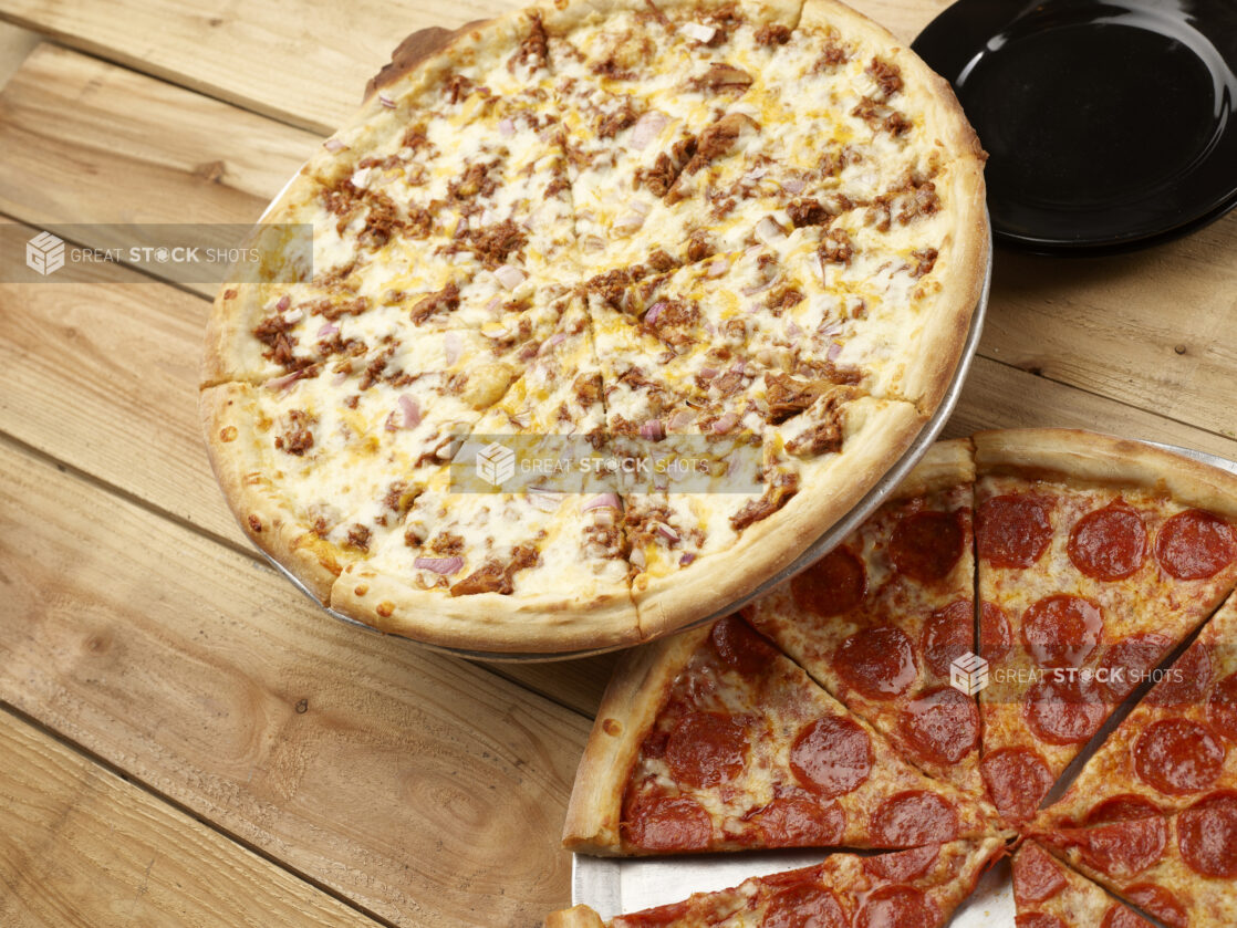 2 Whole pizzas, one with pulled pork and red onions, the other one with pepperoni, both sliced on a wooden background