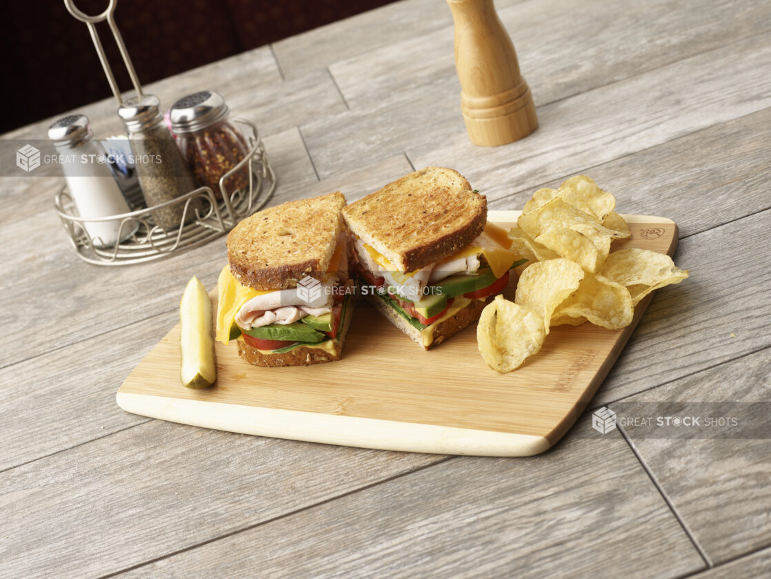 Deli turkey sandwich, cut in half, with avocado, tomato, cheese and honey mustard on multigrain rye on a wooden board with potato chips and slice of pickle, condiments in the background