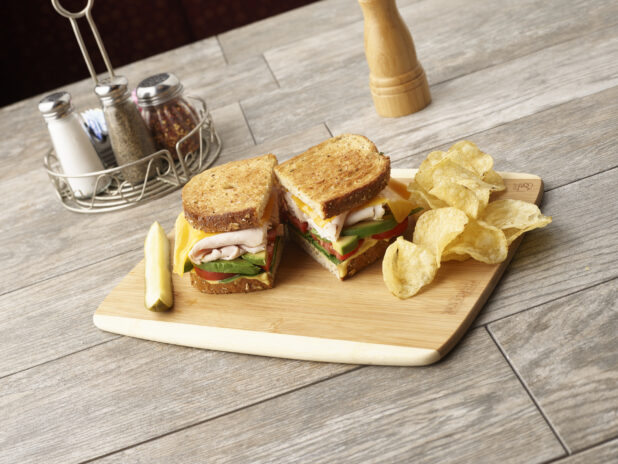 Deli turkey sandwich, cut in half, with avocado, tomato, cheese and honey mustard on multigrain rye on a wooden board with potato chips and slice of pickle, condiments in the background