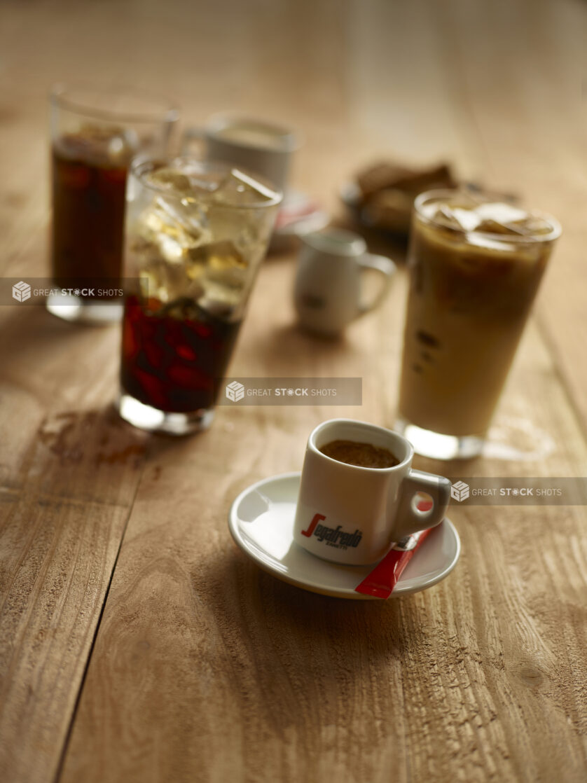 Close up view of an espresso with sugar on the side with iced coffees in the background on a rustic wooden background