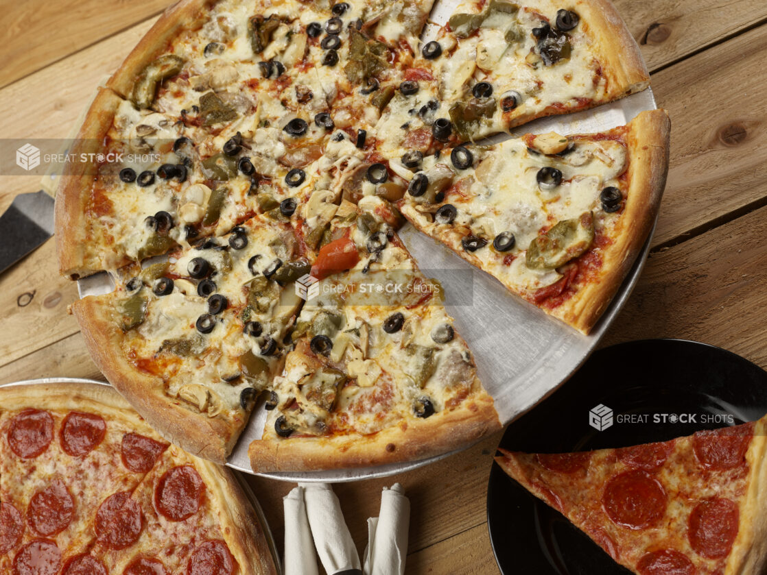 Whole 3 topping pizza, sliced, with black olives, roasted peppers and mushrooms and a pepperoni pizza and a slice all on a wooden background