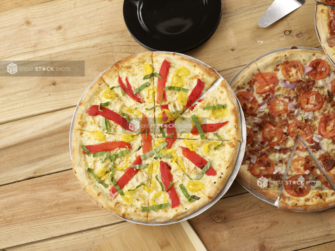 2 California style pizzas, one with roasted peppers, banana peppers and fresh basil, the other with bacon, red onion and tomatoes all on a wood background