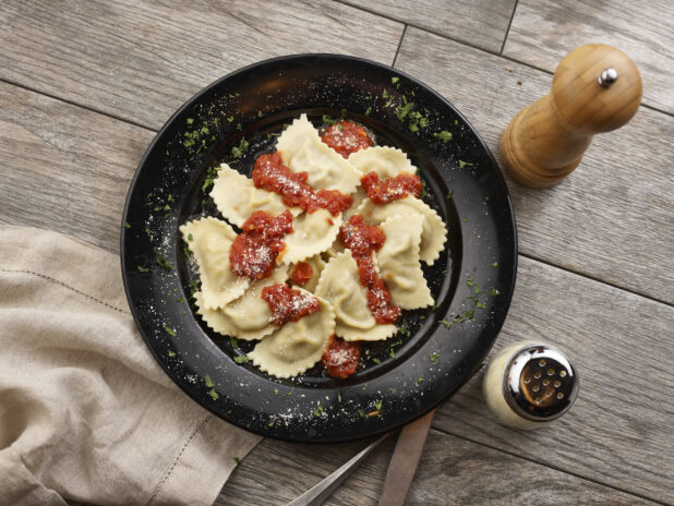 Agnolotti with tomato sauce and parmesan in a black bowl on a grey wooden background, overhead view