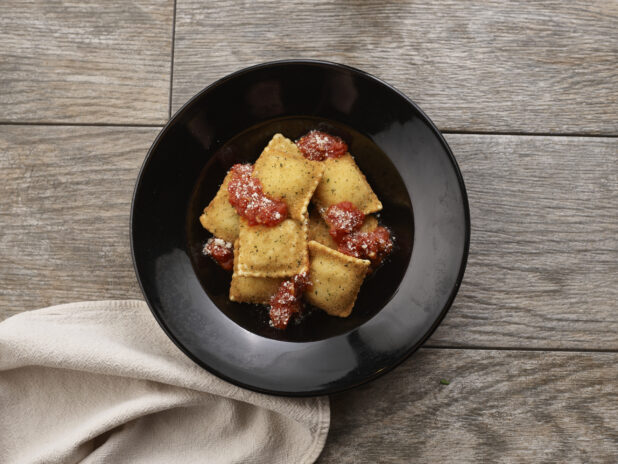 Deep fried ravioli with tomato sauce in a round black bowl on a grey wooden background, overhead view