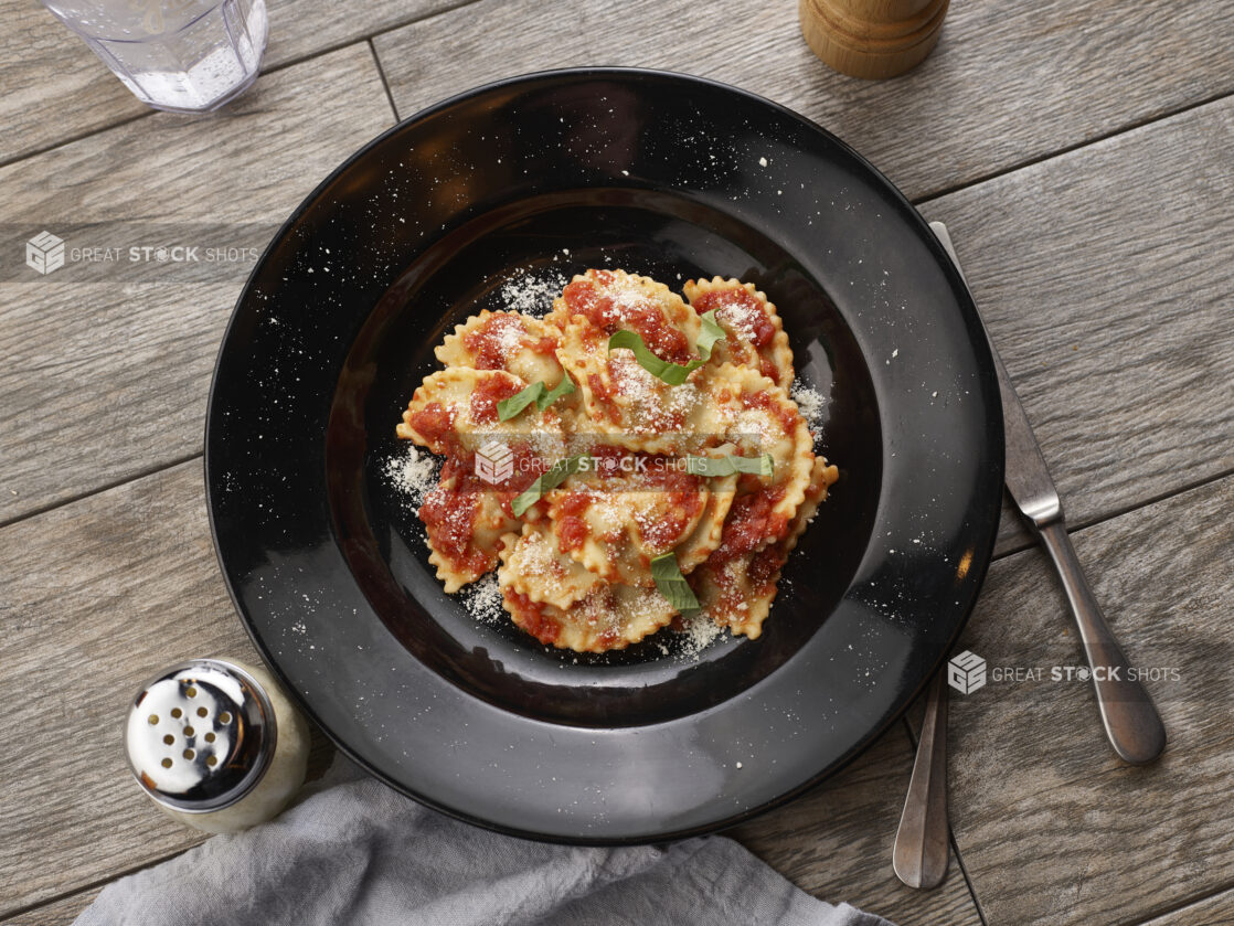 Agnolotti with tomato sauce and fresh basil in a black bowl on a grey wooden background, overhead view