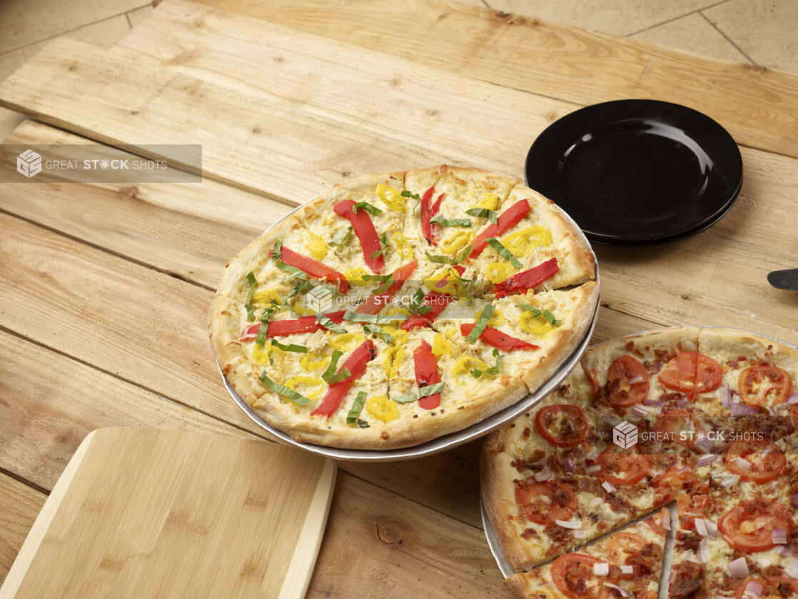 2 whole California-style pizzas, one with banana peppers, roasted bell peppers and fresh basil, the other with bacon, tomatoes and red onion on a wooden background