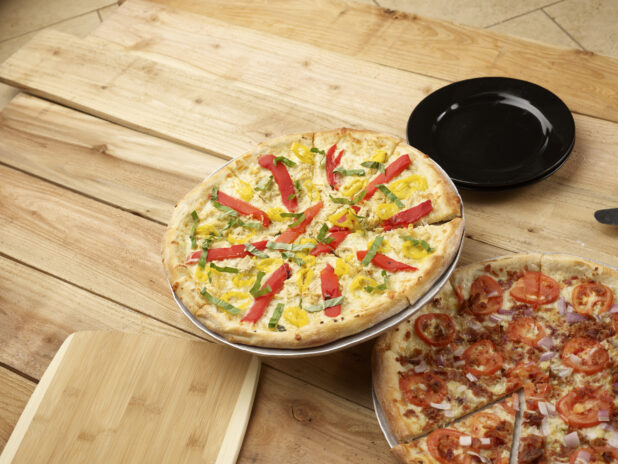 2 whole California-style pizzas, one with banana peppers, roasted bell peppers and fresh basil, the other with bacon, tomatoes and red onion on a wooden background