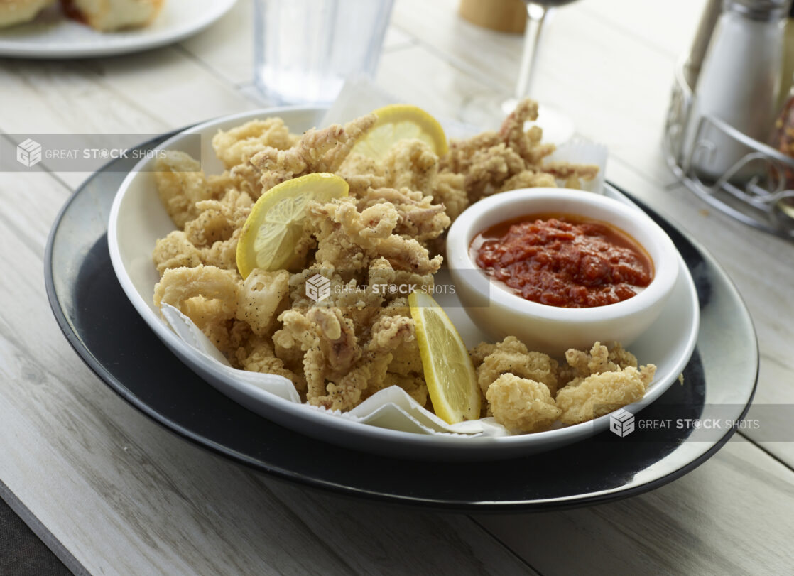 Deep fried calamari with lemon slices and a ramekin of seafood sauce in a white bowl on a dark plate in a dining setting, close up view