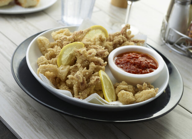 Deep fried calamari with lemon slices and a ramekin of seafood sauce in a white bowl on a dark plate in a dining setting, close up view