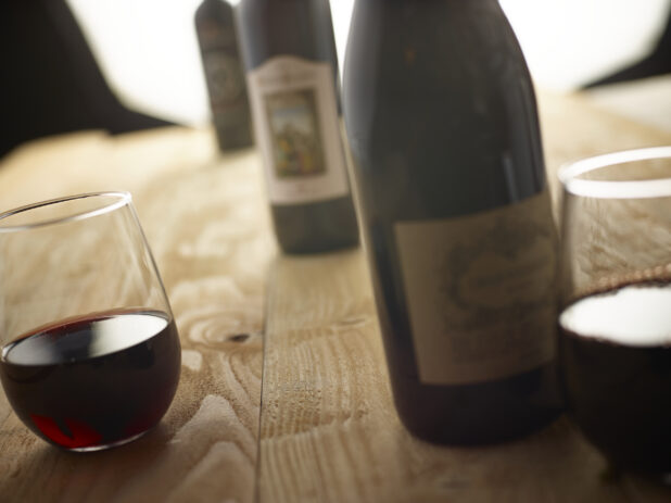 Bottles of red wine and stemless wine glasses with red wine on an unfinished wooden tabletop