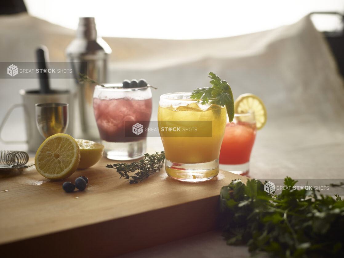 Old fashioned glasses with various mixed drinks on a wooden board with fresh lemon, blueberries and fresh herbs surrounding, with mixing utensils in the background