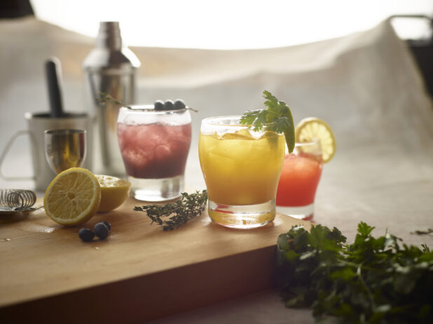 Old fashioned glasses with various mixed drinks on a wooden board with fresh lemon, blueberries and fresh herbs surrounding, with mixing utensils in the background