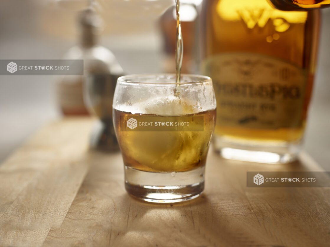 Whiskey/old fashioned glass with round ice cube filled with an amber alcohol with a bottle in the background on a wooden tabletop