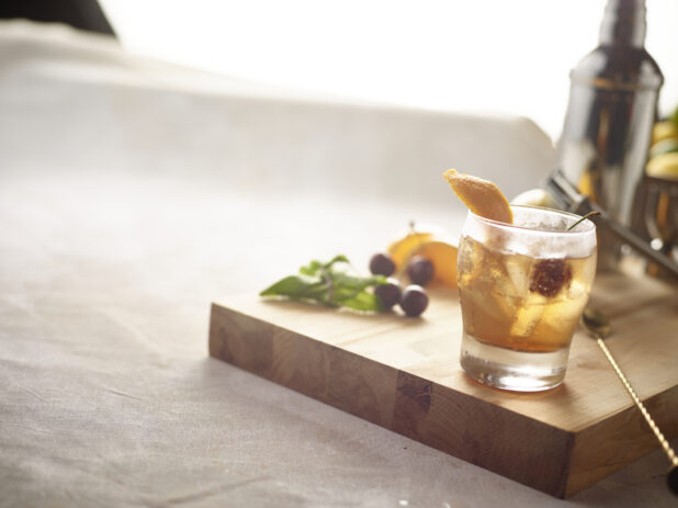 Cocktail in a whiskey/old fashioned glass with fresh orange and cherry garnish on a wooden board with mint cherries and orange garnish on the side