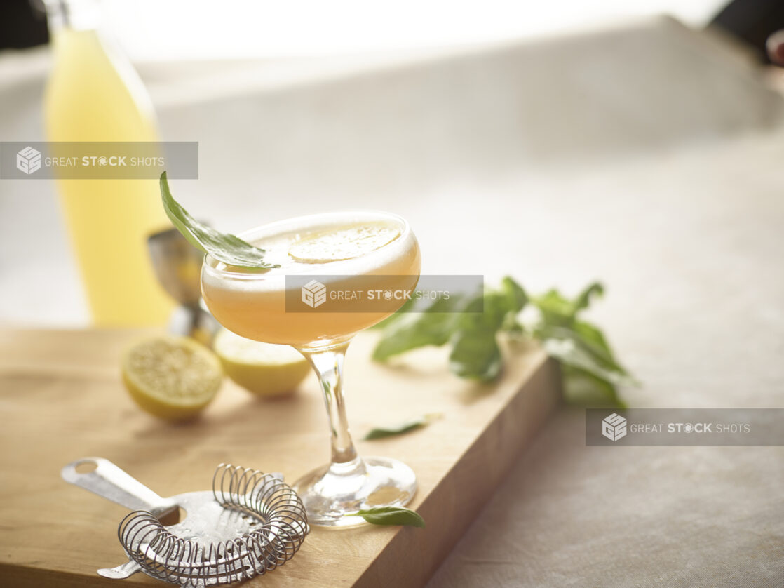 Cocktail in a coupe/champagne glass on a wooden board with strainer, lemon, basil surrounding