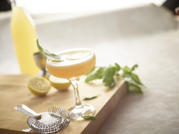 Cocktail in a coupe/champagne glass on a wooden board with strainer, lemon, basil surrounding