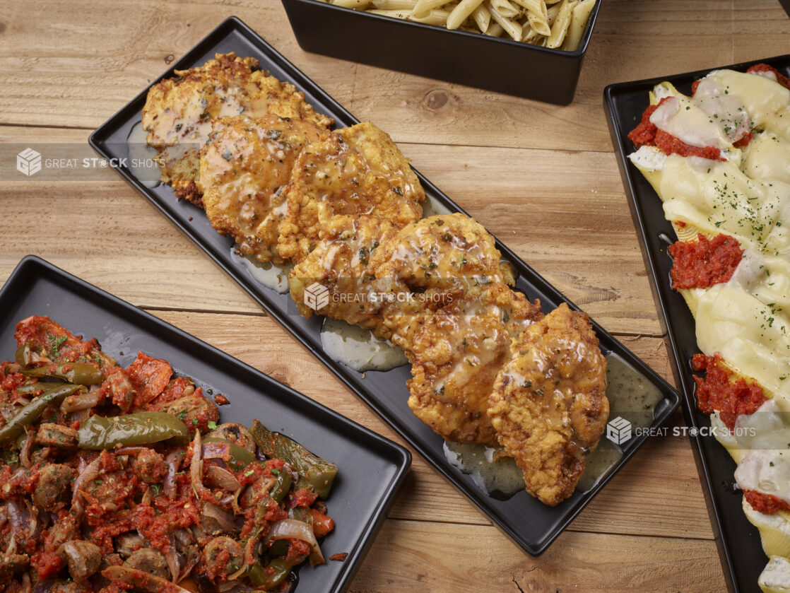 Chicken scallopini, sauteed bell peppers and onions in tomato sauce, stuffed pasta shells and penne pasta all on black platters on a wooden background