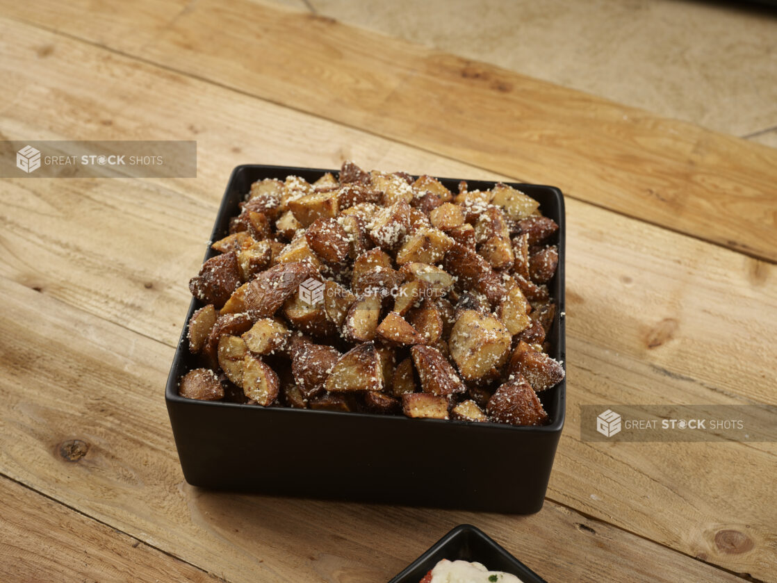 Roasted parmesan potatoes in a square black bowl on a wooden background