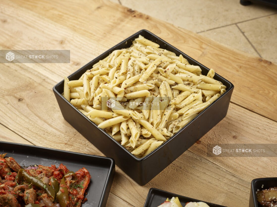 Penne pasta with olive oil and seasoning in a square black bowl on a wooden background