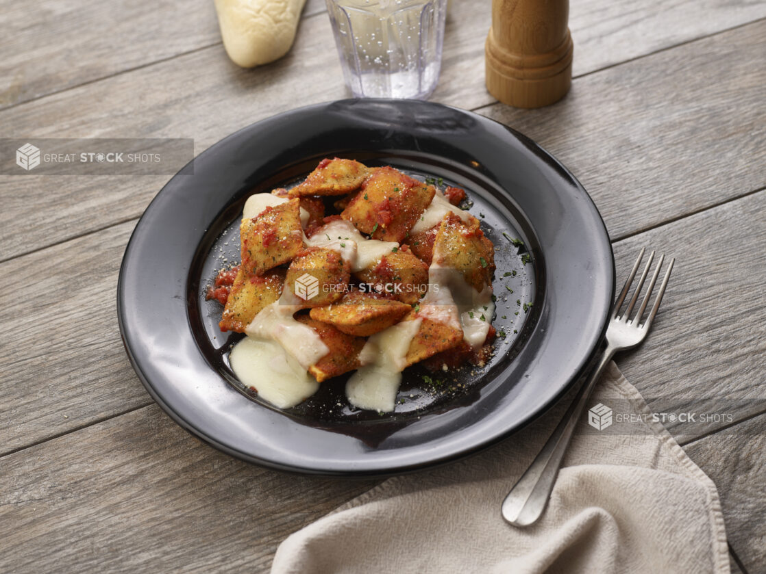 Deep fried ravioli with melted mozzarella and tomato sauce in a round black bowl on a grey wooden background