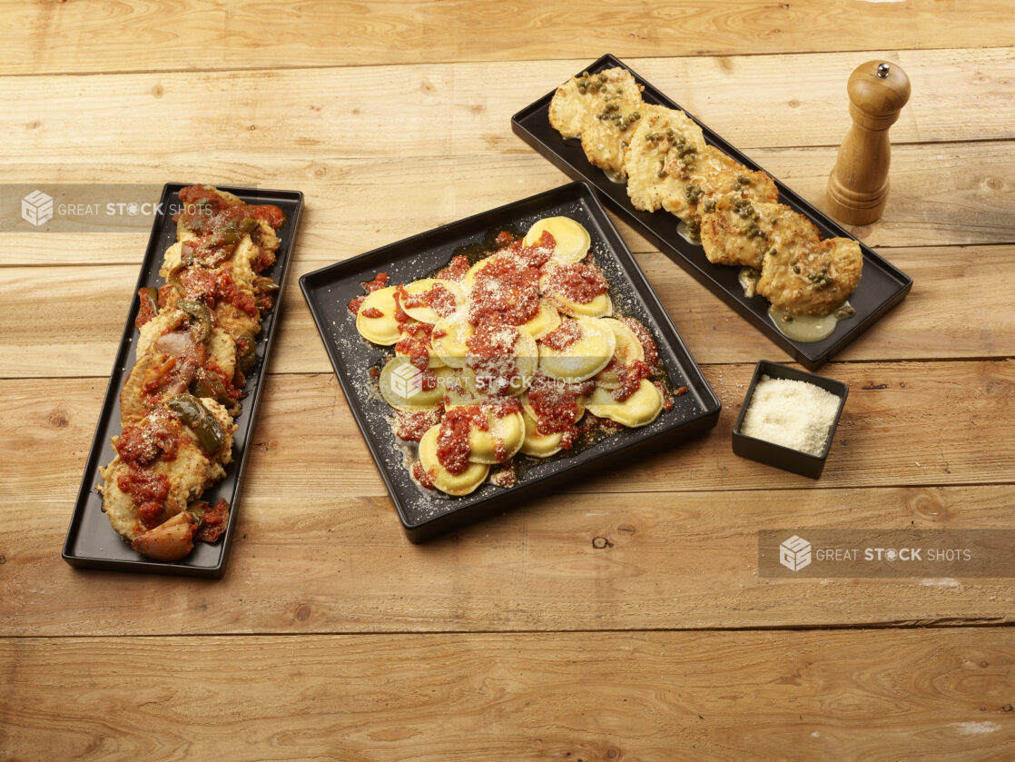 3 kinds of ravioli on black platters with a small ramekin of parmesan cheese and a pepper mill on a wooden background