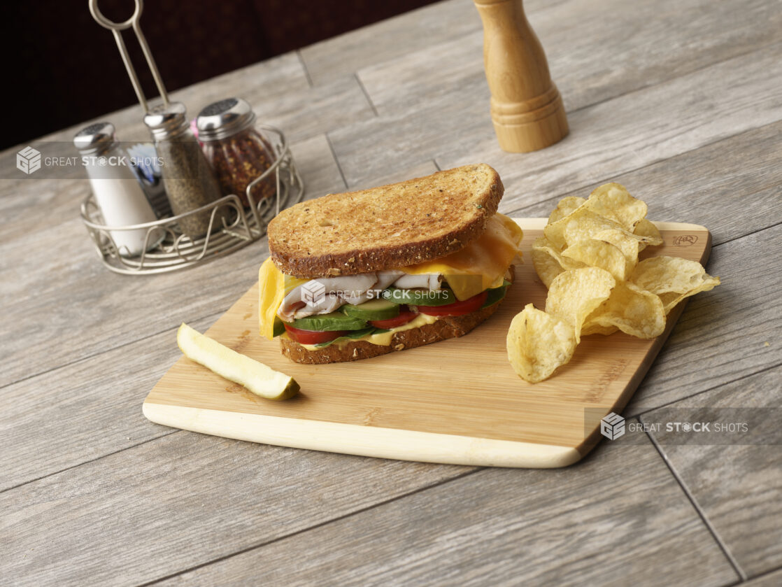 Deli turkey sandwich with avocado, tomato, cheese and honey mustard on multigrain rye on a wooden board with potato chips and slice of pickle, condiments in the background