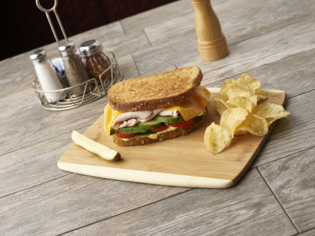 Deli turkey sandwich with avocado, tomato, cheese and honey mustard on multigrain rye on a wooden board with potato chips and slice of pickle, condiments in the background
