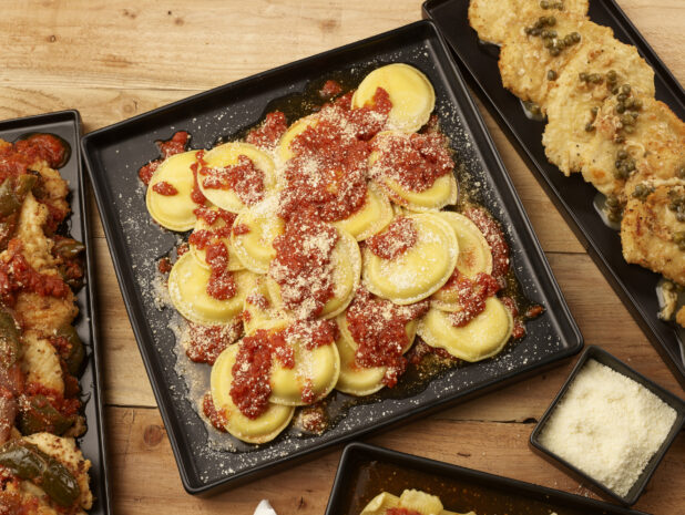 Mezzalune ravioli with fresh tomato sauce and grated parmesan surrounded by other Italian dishes, all on black square and rectangular platters