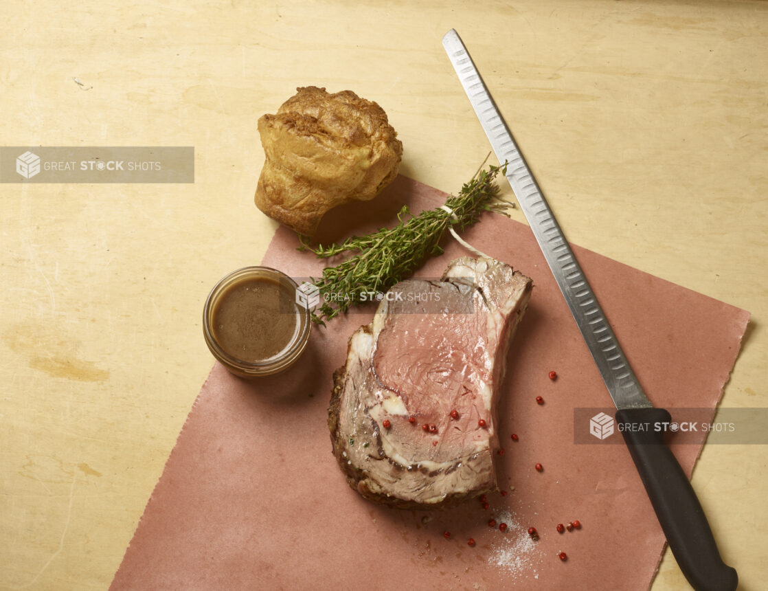 Slice of roast beef with a side of gravy in a mason jar, Yorkshire pudding, fresh thyme and a carving knife with some salt and pink peppercorns scattered around on butchers paper on a wooden background