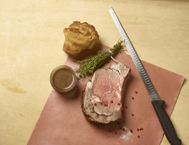 Slice of roast beef with a side of gravy in a mason jar, Yorkshire pudding, fresh thyme and a carving knife with some salt and pink peppercorns scattered around on butchers paper on a wooden background