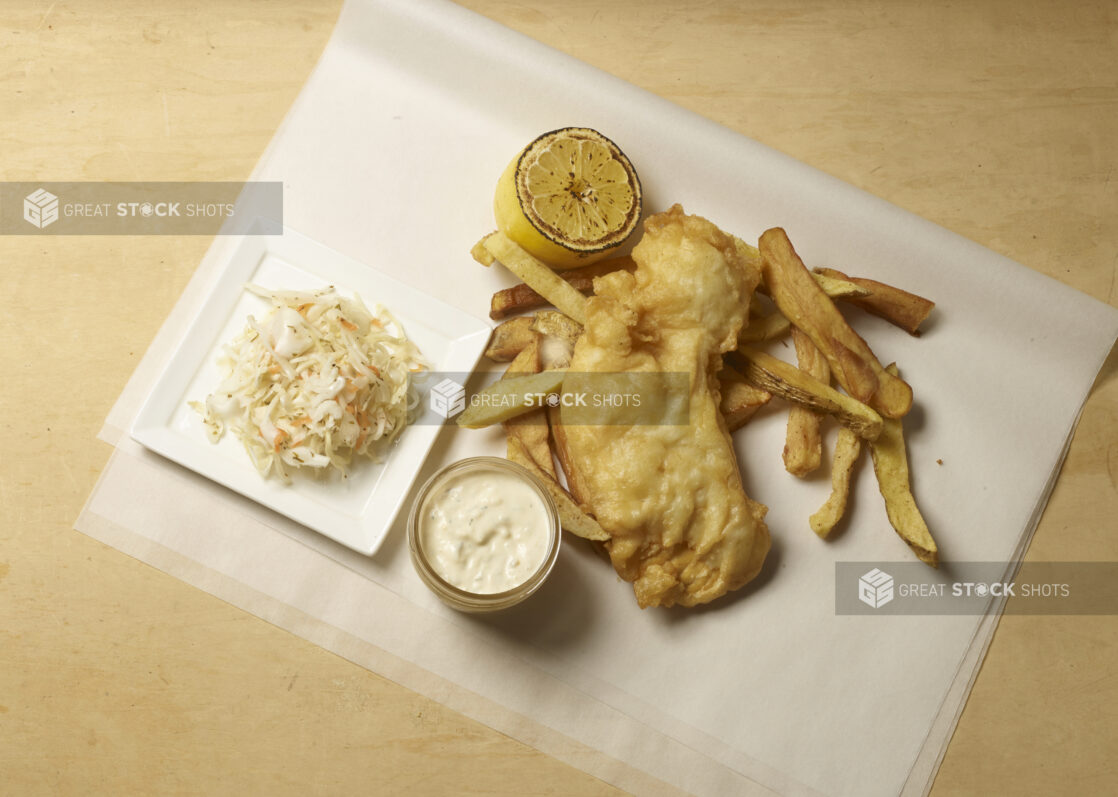 Fish and chips on parchment paper with a side of coleslaw, grilled lemon and a side of tartar sauce on a wooden background