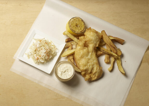 Fish and chips on parchment paper with a side of coleslaw, grilled lemon and a side of tartar sauce on a wooden background