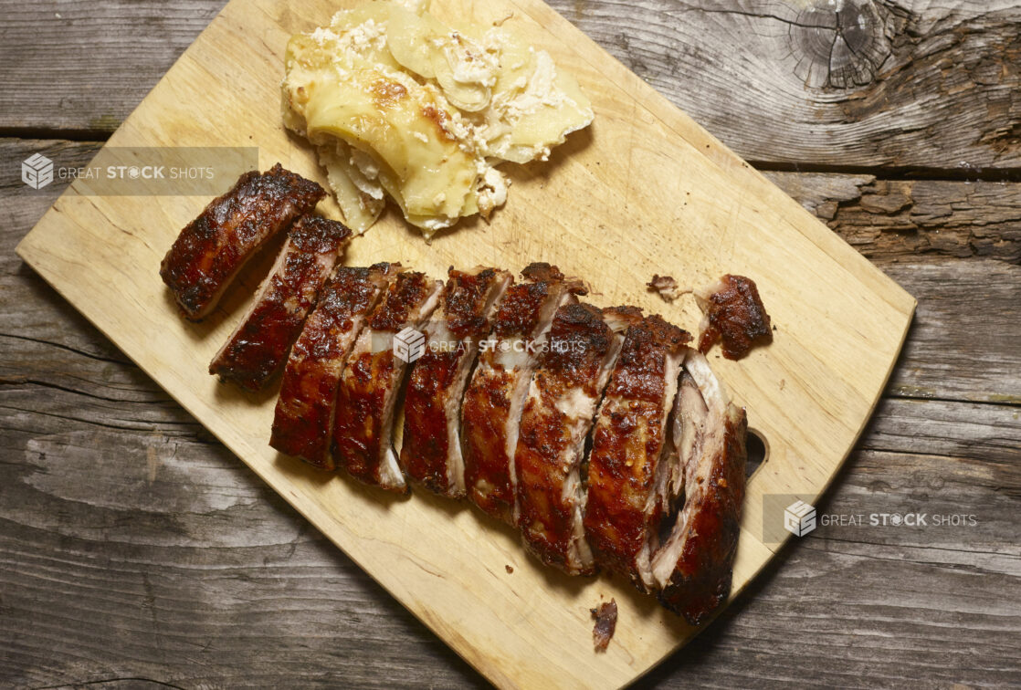 Full rack of BBQ ribs with a side of scalloped potatoes on a wooden cutting board on a rustic wooden background