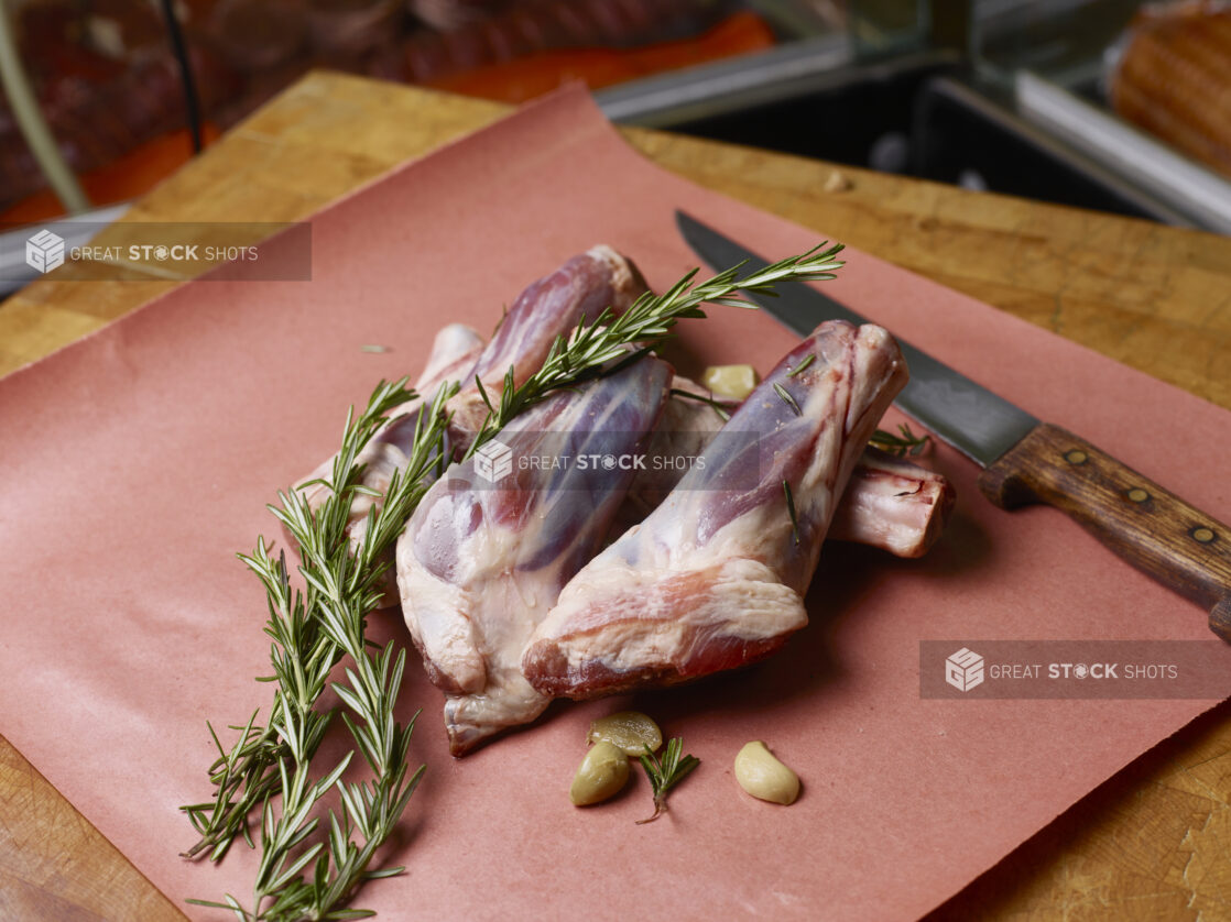 Uncooked lamb shanks on butcher's paper with garlic cloves and fresh rosemary with a knife on a butchers block