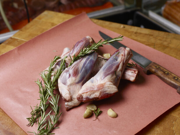 Uncooked lamb shanks on butcher's paper with garlic cloves and fresh rosemary with a knife on a butchers block