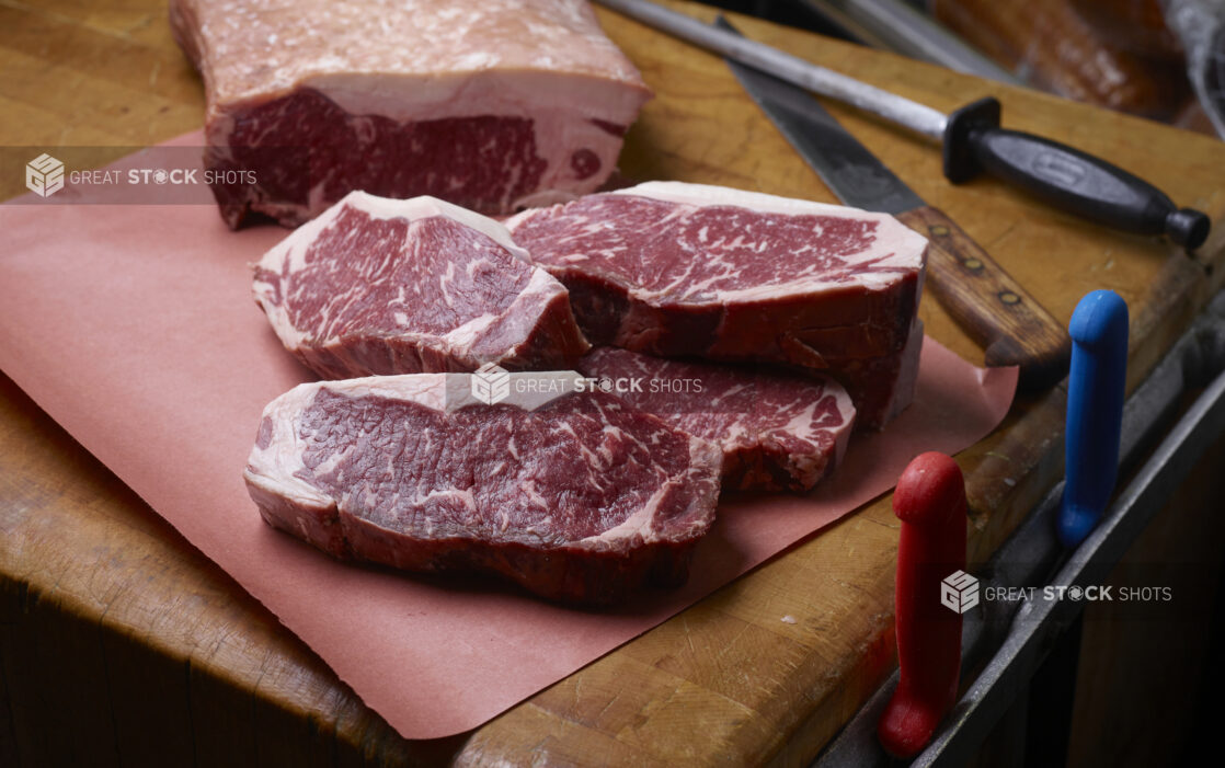 Raw uncooked whole strip roast with a few steaks cut from it on butcher paper on a butcher's block, knife and sharpening steel in background