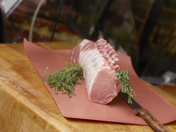 Whole pork rib rack roast, uncooked, on butchers paper with fresh thyme and a boning knife on a butchers block