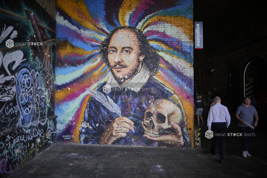 Mural of William Shakespeare holding a skull and a quill on a wall in London, England with people walking by the mural