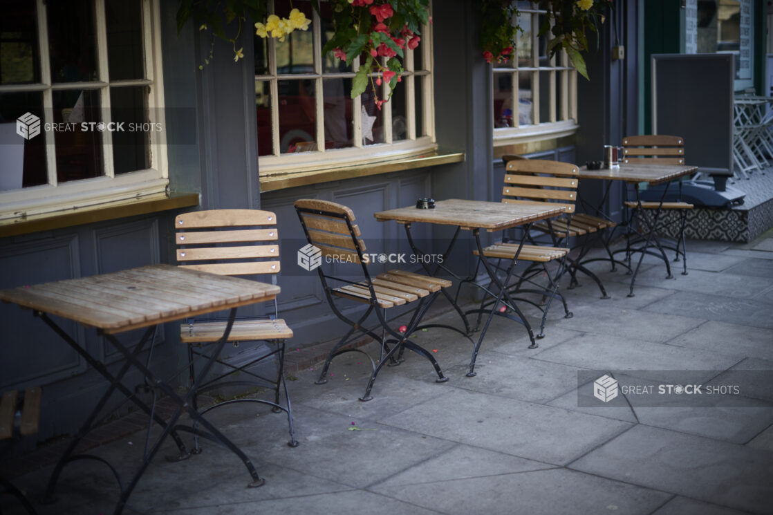 Outdoor cafe setting with wooden slatted chairs and tables with wrought iron legs, beside windows in hanging flowers