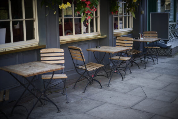 Outdoor cafe setting with wooden slatted chairs and tables with wrought iron legs, beside windows in hanging flowers